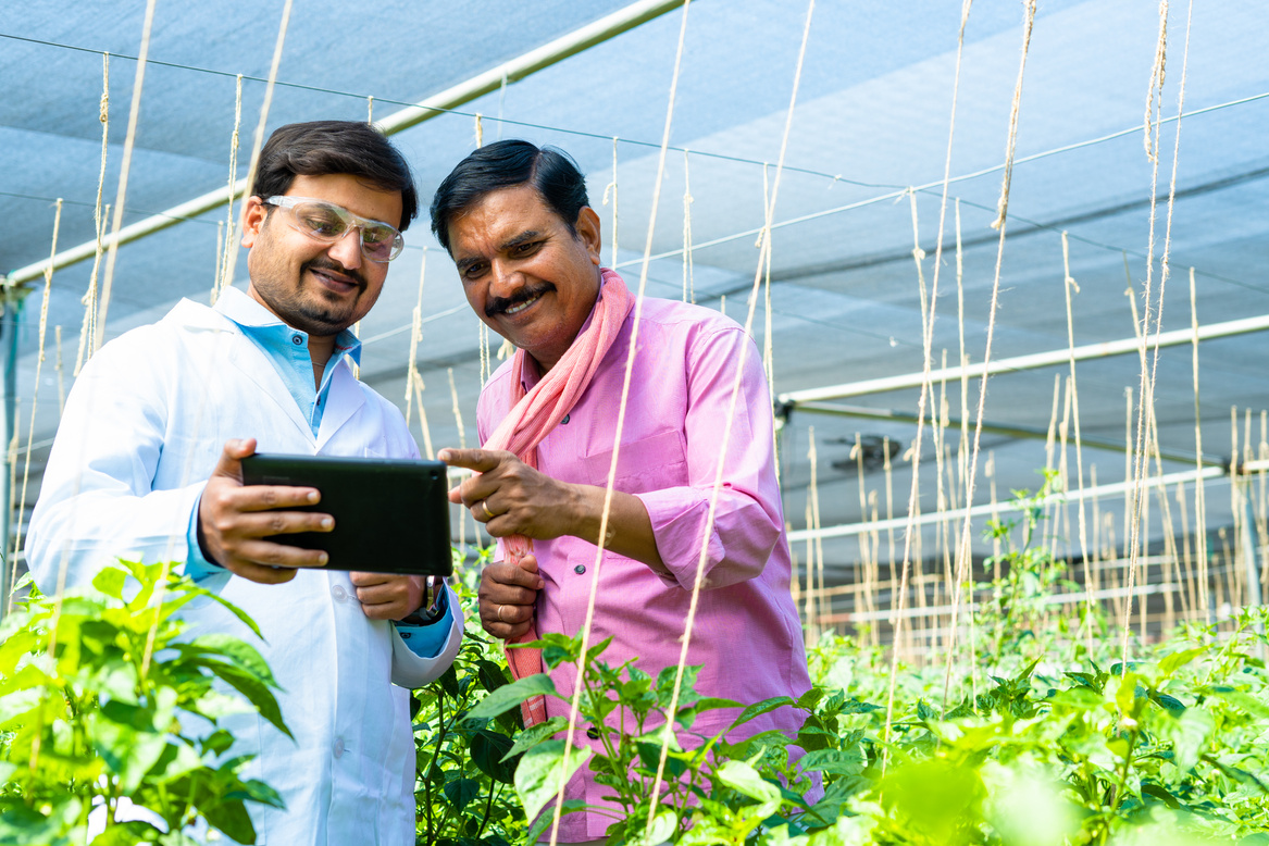 Indian happy agro scientist with farmer checking tablet about plant growth at greenhouse - concept of expert assistance, guidance and biotechnology